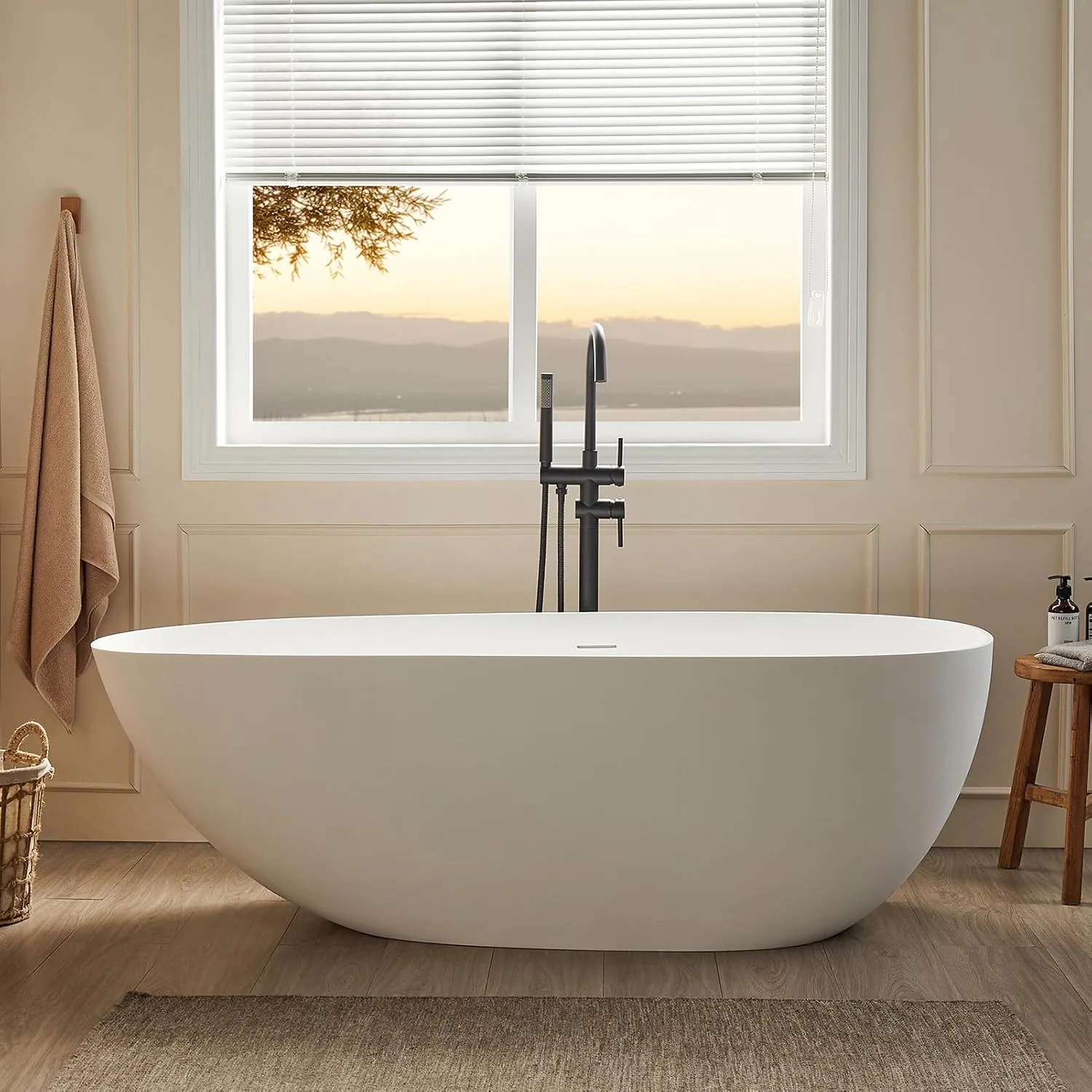 Matte white freestanding soaking tub in a warm neutral bathroom with wood flooring, soft towels, black faucet, window light, and calm spa-like styling.